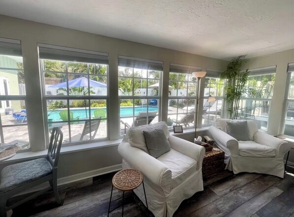 A sunroom with large windows, two white sofas, a wicker chair, and a small table.