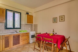A kitchen with wooden cabinets, a tiled backsplash, a window with curtains, a dining table with a red tablecloth, and two framed pictures on the wall. - Analipsi Villas Beach Front (Andravida-Kyllini)