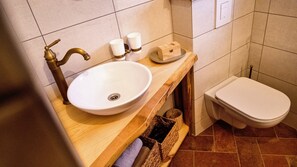 Un baño con tocador de madera, un lavabo blanco, dos tazas blancas, un pequeño cofre de madera y un inodoro blanco.