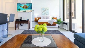 A modern hotel room with a bed, a desk, a chair, a coffee table with a vase of flowers, and a painting on the wall.