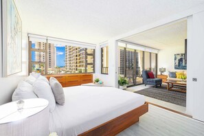A modern bedroom with a large bed, a wooden dresser, and a view of a cityscape through large windows. - 1BR near Waikiki Beach w/ pool & hot tub (Honolulu)