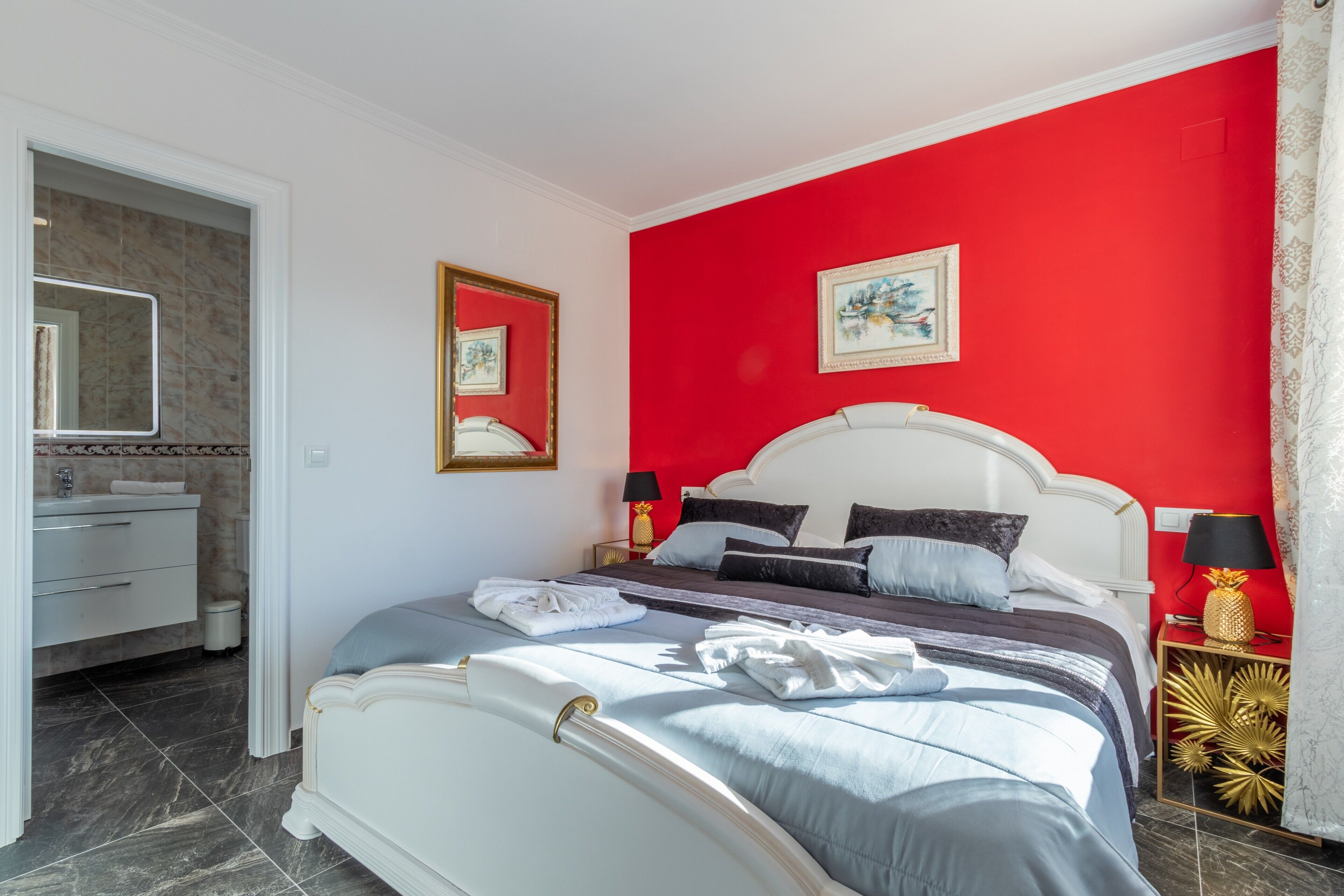 A hotel room with a bed, a nightstand, a lamp, a mirror, a painting, and a bathroom with a sink and a towel rack.