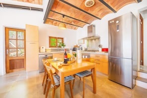 A modern kitchen with a wooden dining table, chairs, a stainless steel refrigerator, and a window with a view of greenery.