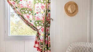 Ein Zimmer mit Fenster, einem Vorhang mit Blumenmuster, einem Korbstuhl und einem Strohhut an der Wand.
