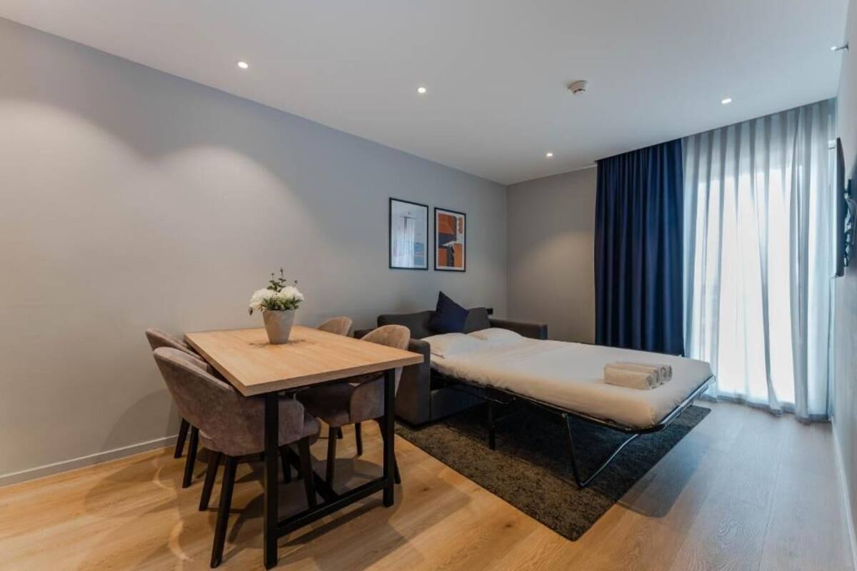 A modern room with a wooden dining table, chairs, a sofa, a bed, and a large window with curtains.