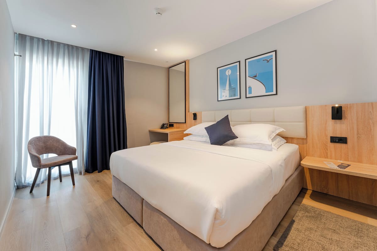 A modern hotel room with a large bed, a bedside table, a chair, and a large window with curtains.