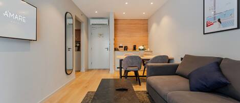 A modern hotel room with a sofa, a coffee table, a dining area with chairs, and a kitchen in the background.