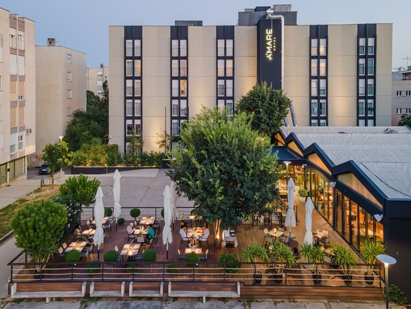 Un hotel con zonas de comedor al aire libre, un gran árbol y una fachada de edificio moderna.