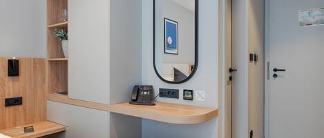 Una habitación de hotel moderna con un escritorio de madera, un espejo redondo, un teléfono en la mesita de noche y un pequeño taburete.