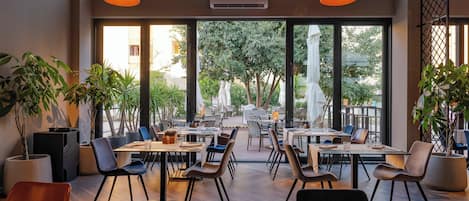 Un comedor interior con mesas y sillas, ventanas amplias y plantas en macetas.