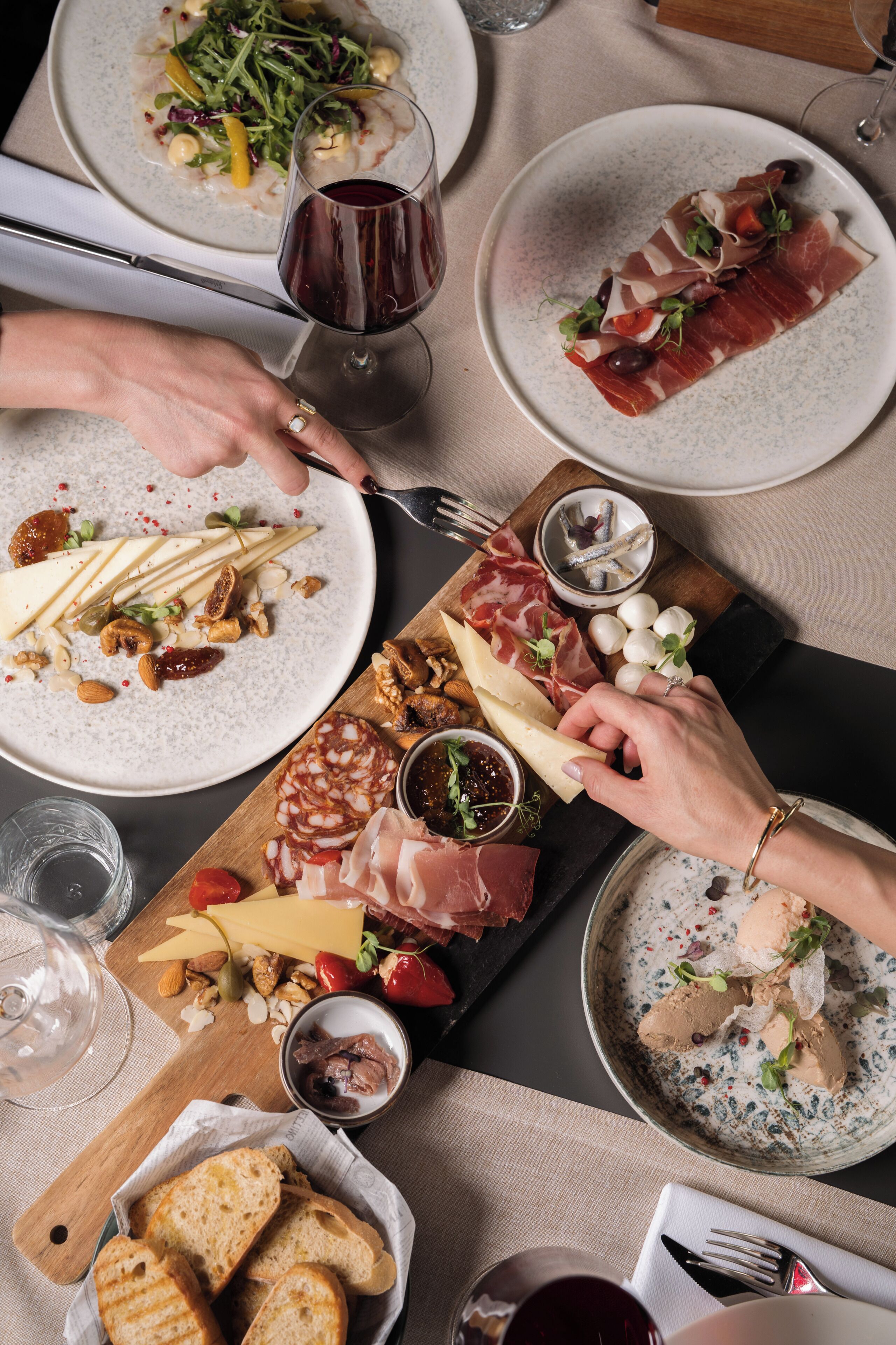 Una mesa servida con varios quesos, embutidos y pan, acompañada de copas de vino tinto.
