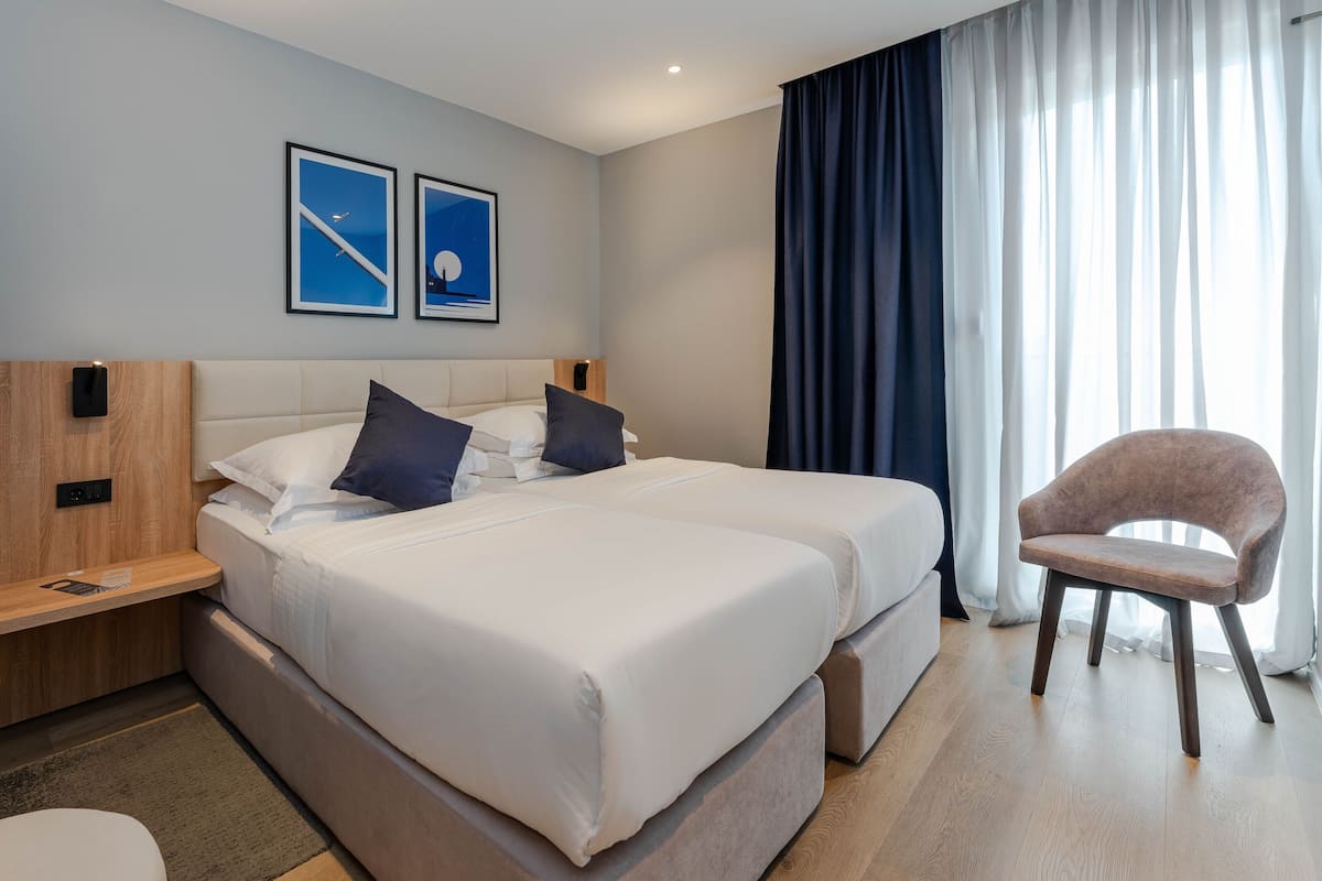 A hotel room with two beds, a wooden nightstand, a chair, and a large window with curtains.