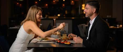 A couple dining at a table in a dimly lit restaurant with a view of the cityscape.