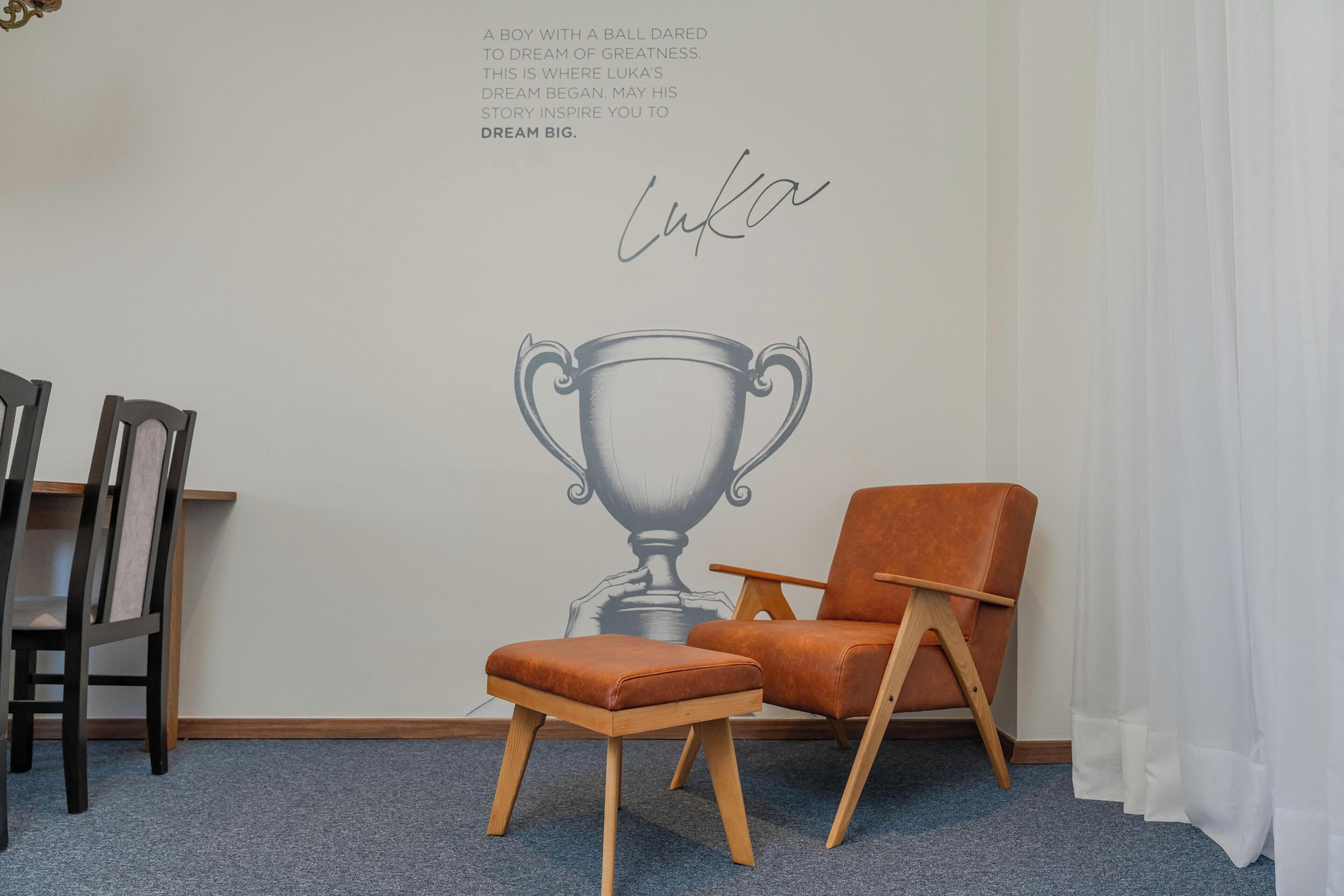 Una habitación con un mural en la pared que muestra un trofeo, una silla de madera, un taburete acolchado y una cortina.