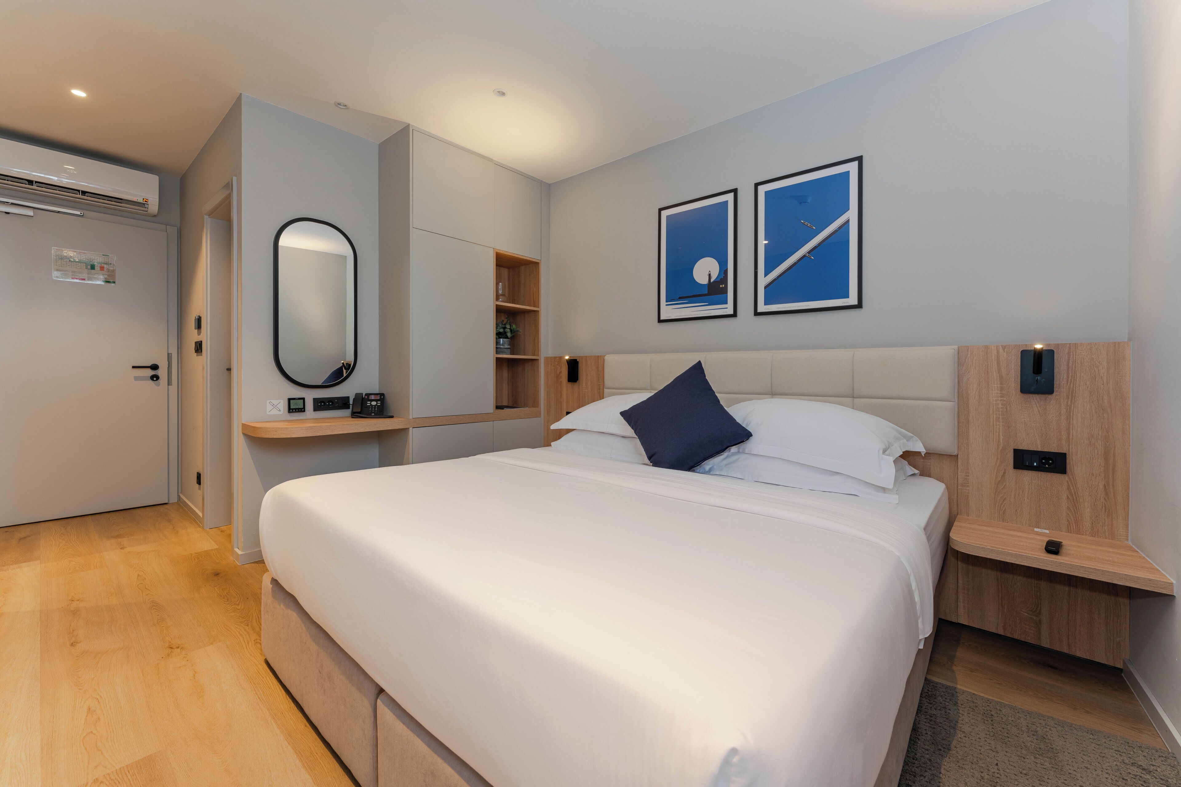 A modern hotel room with a large bed, a built-in wardrobe, a bedside table, a wall-mounted mirror, and two framed pictures.