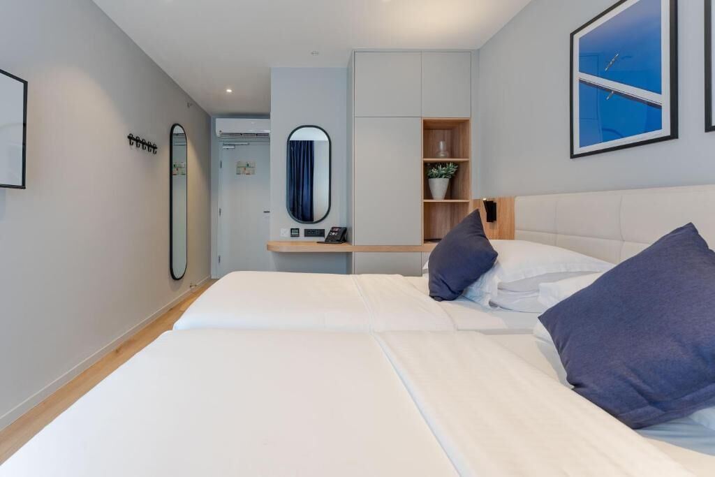 Un dormitorio moderno con dos camas, un espejo, una estantería con una planta y una obra de arte azul en la pared.
