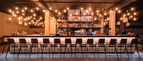 A bar area with a long counter, multiple hanging light fixtures, and a variety of bottles and glasses.