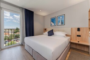 Habitación de hotel moderna con una cama grande, una mesita de noche de madera y vistas a un paisaje urbano a través de un ventanal amplio.