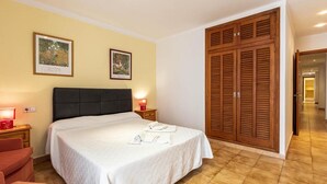A hotel room with a bed, two bedside tables with lamps, a wooden wardrobe, and two framed pictures on the wall. - Holiday Apartment 'Daniela' next to the Beach with Sea View, A/C, Balcony & Wi-Fi (Pollenca)