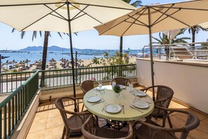 Une terrasse dotée de chaises et d’une table en osier, aménagée pour les repas, et offrant une vue sur une plage où l’on aperçoit des bateaux et des palmiers.