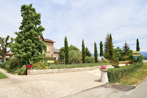 A residential area with a stone house, a gravel driveway, and a variety of trees and shrubs. - Holiday Home 'Gîte Les Galets Chantants' with Mountain View, Private Terrace and Wi-Fi (Saint-Hilaire-Du-Rosier)