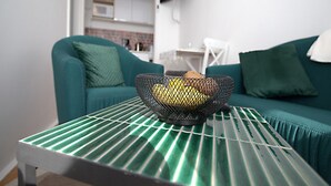 Una sala de estar moderna con un sofá verde, una mesita con una cesta de alambre, y una cocina al fondo.