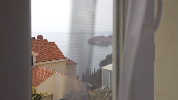 Vista a través de una ventana con cortina, mostrando un paisaje costero con edificios y una formación rocosa destacada.