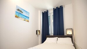 Un dormitorio con una cama, un cuadro de una playa, una ventana con cortinas y una mesita de noche con una lámpara.