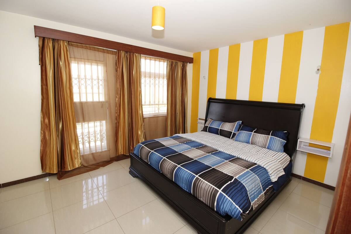 A bedroom with a large bed, curtains, a window, and a yellow and white striped wall.