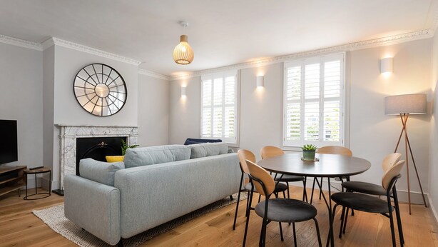 A modern living room with a grey sofa, a fireplace, a dining table with chairs, and a large wall clock.