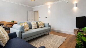 A modern living room with a grey sofa, patterned cushions, a patterned rug, a wooden TV stand, and a hanging light fixture.