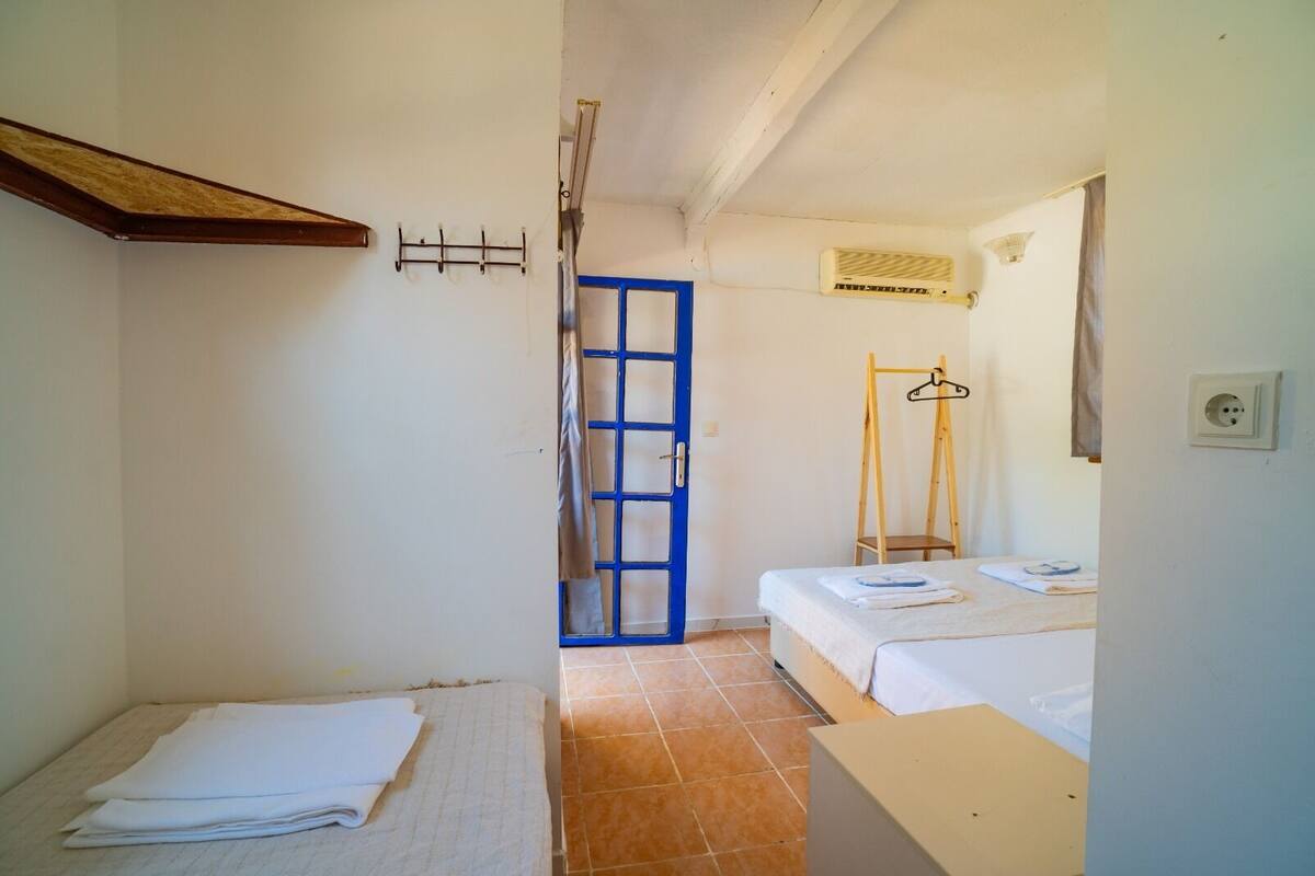 A narrow room with two beds, a blue door, a wooden shelf, and a hanging rack.