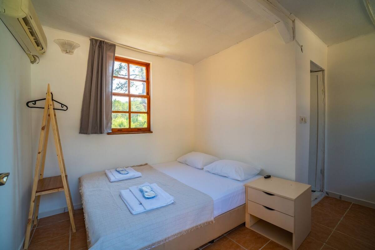 A bedroom with a bed, a bedside table, a wooden ladder, a window with curtains, and a door.