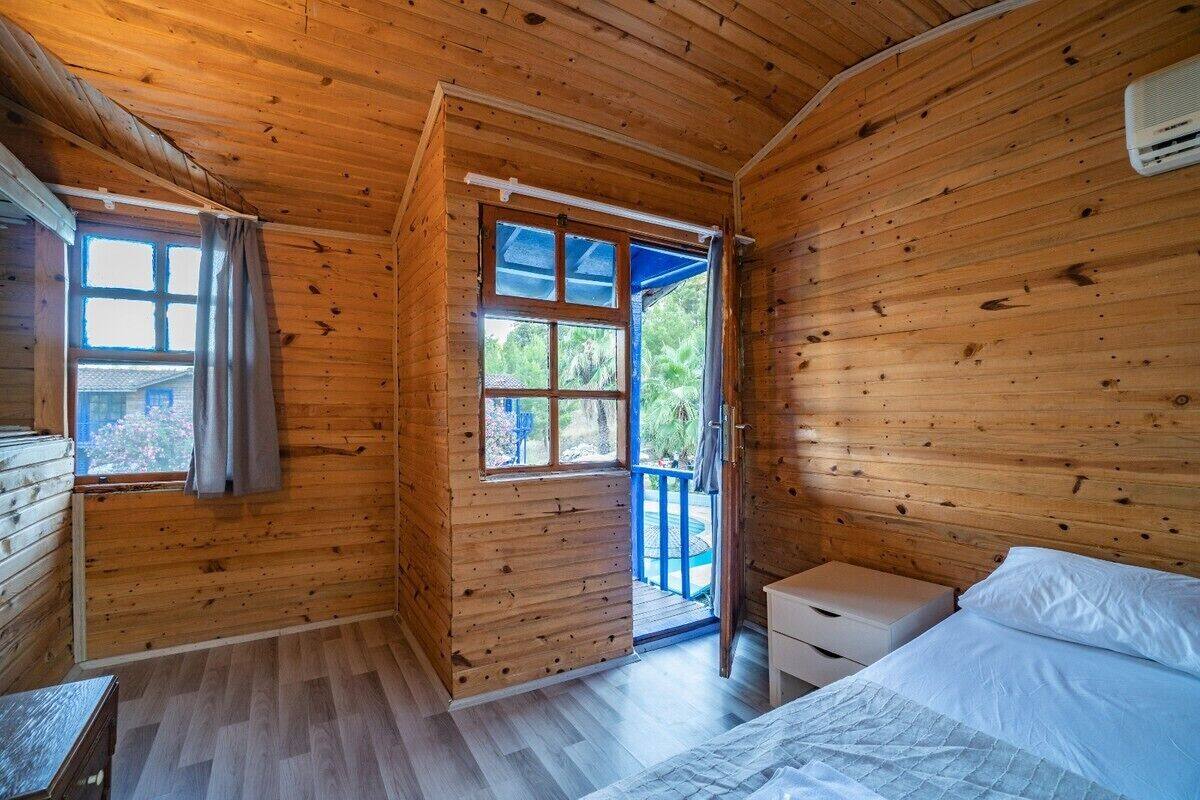 A wooden cabin room with a bed, a small table, and a window with curtains.