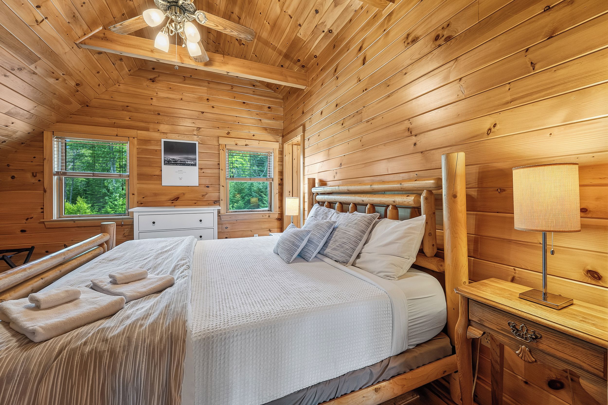 Une chambre dans une cabane en bois, avec un lit, une table de chevet, une commode et un ventilateur de plafond.