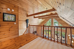 A wooden interior with a staircase leading to an upper level, a framed painting on the wall, and a view of greenery outside.