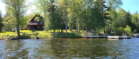 En stuga vid sjön med en brygga och en liten pir, omgiven av frodig grönska och träd.