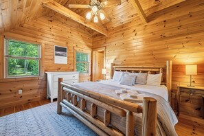 A wooden cabin bedroom with a bed, bedside table, lamp, and a view of the forest through the window.
