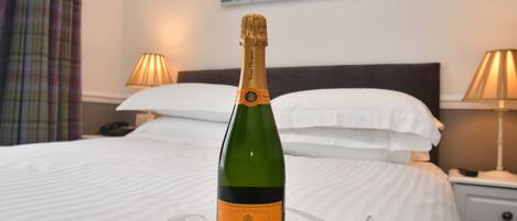 A bottle of Veuve Clicquot Brut champagne on a clear glass tray on a bed with white linens, two bedside lamps, and a painting on the wall.