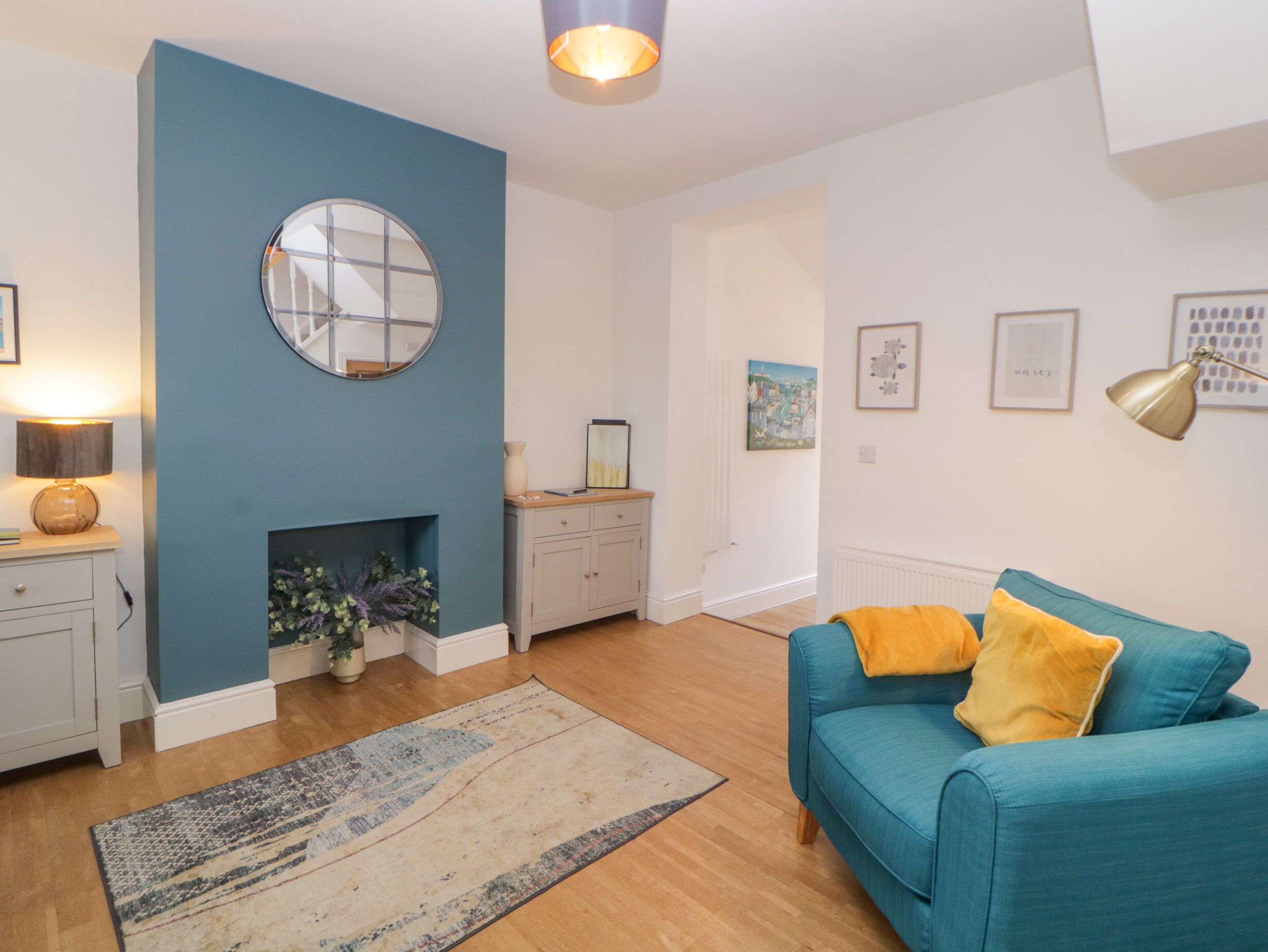 A modern living room with a blue accent wall, a round mirror, a fireplace, a teal sofa with yellow cushions, and a wooden floor.