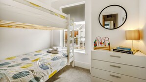 Una cama litera con colcha de estampado de hojas, una mesita de noche con lámpara y libros, y un espejo en la pared.