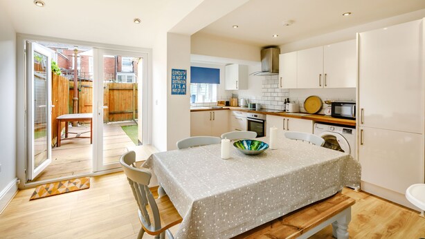 Una cocina moderna con armarios blancos, una mesa de comedor con mantel blanco y vista al jardín trasero a través de puertas de vidrio.