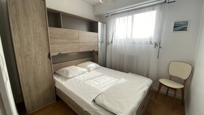 Un dormitori amb un llit de fusta, una cadira, una finestra amb cortines translúcides i una làmpada penjant.