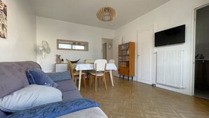 Un saló amb un sofà, taula de dinar, cadirees, una prestatgeria, un televisor i un radiador.