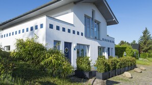 Ett modernt hus i två våningar med vit fasad, en blå dörr och en liten trädgård med buskar och ett träd.