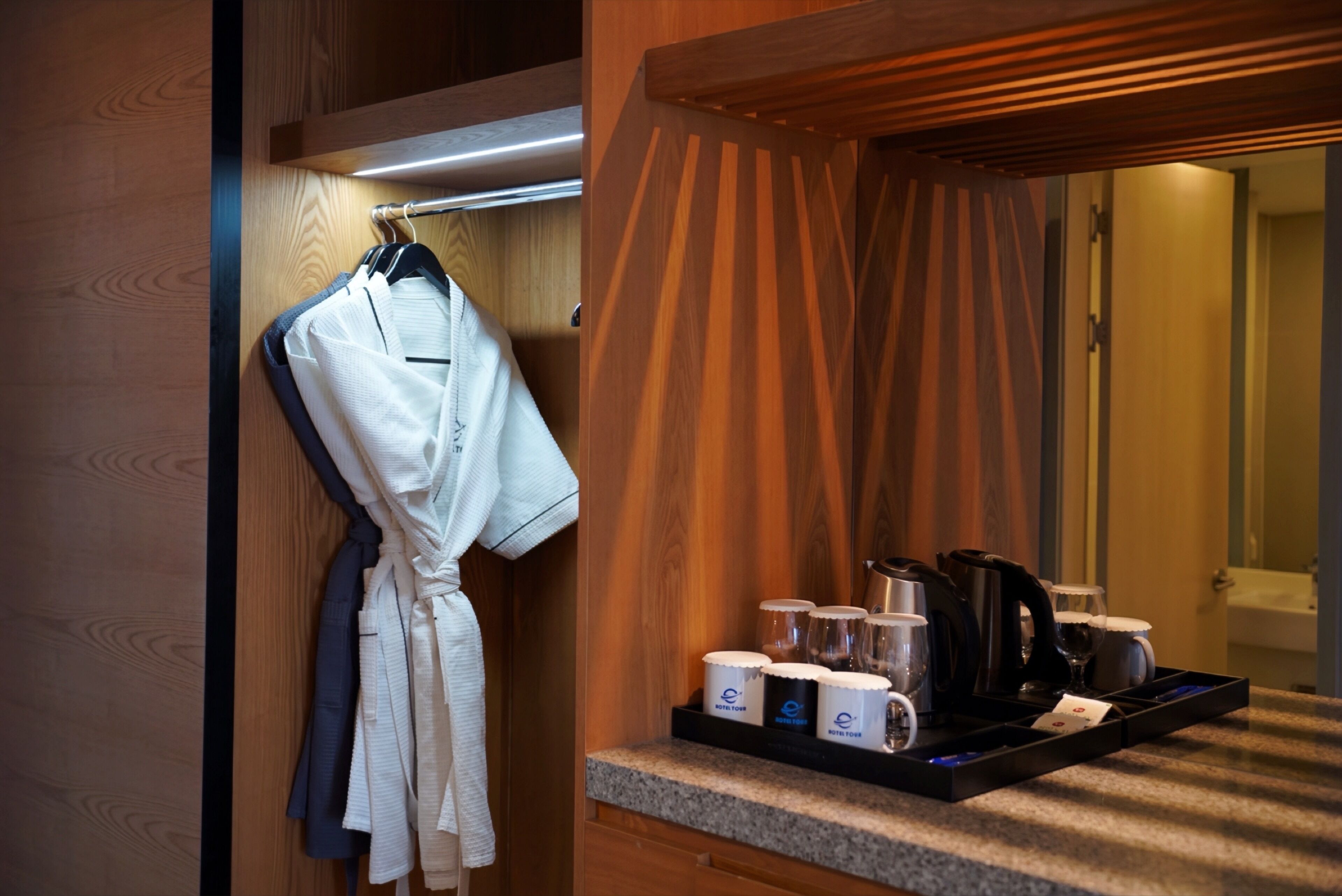 A hotel bathroom with a wooden vanity, a towel rack with robes, and a tray with mugs and a coffee maker.