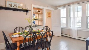 A dining area with a wooden table set for four, surrounded by black chairs. A framed picture hangs above the table, and a small shelf with decorative items is visible on the wall. - Walk to Restaurants Shopping & Harbor! (Wellfleet)