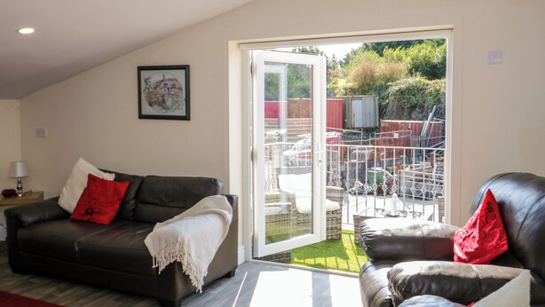 A living room with a brown leather sofa, a red throw blanket, and a white throw blanket. There is a glass door leading to an outdoor area with a patio and a view of a garden. - The Apartment at Plas Yr Eithin (Amlwch)