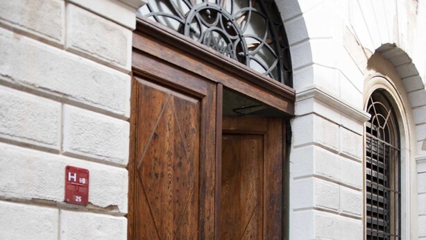 Une grande porte en bois, surmontée d’une grille métallique, est intégrée à un bâtiment aux murs en briques blanches et à la base en pierre.