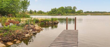 A wooden dock extending into calm water, surrounded by greenery and a distant boat.